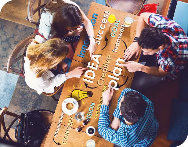 Vista aérea de un grupo de personas reunidas alrededor de una mesa con las palabras "idea", "creativo", "plan" y "concepto" escritas. Representa una sesión de lluvia de ideas o planificación estratégica.